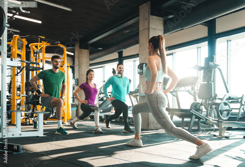 Group of people exercising in a health club. Cheerful young athletes doing lunges in gym under supervision of their attractive coach. Female fitness instructor stretching with a group of three people.