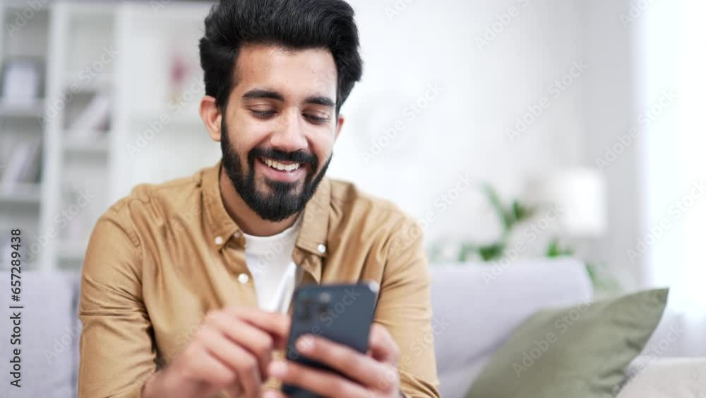 Close up. Young bearded man using smartphone sitting on sofa in living room at home. Smiling handsome male browses social networks, goes shopping online, chats with a friend, writes or reads messages Close up. Young bearded man using smartphone sitting on sofa in living room at home. Smiling handsome male browses social networks, goes shopping online, chats with a friend, writes or reads messages