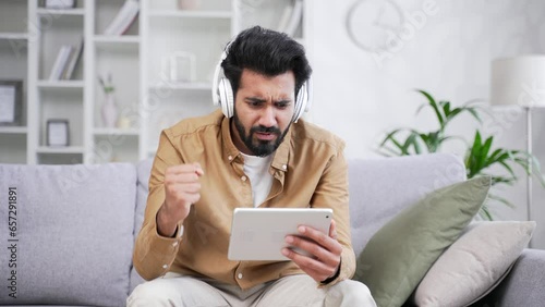 Excited young handsome bearded man in wireless headphones watching sports match competition on a tablet while sitting on sofa in living room at home. Happy male emotionally cheering for favorite team