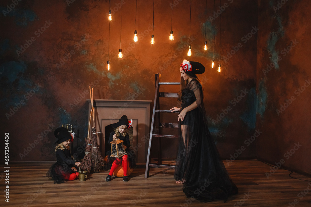 Girls in witch costumes are sitting near the fireplace making a potion, their mother is standing nearby. Girls in witch costumes are sitting near the fireplace making a potion, their mother is standing nearby.