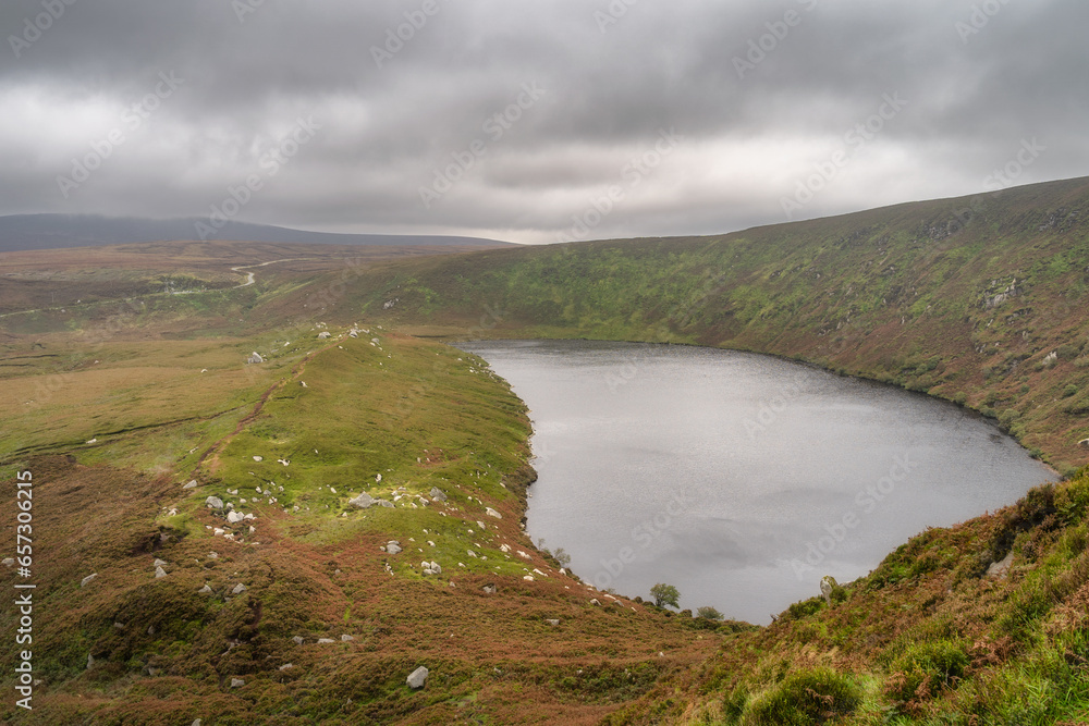 Lake, Lough Bray with mountain trail on the left and cliffs on right, covered in moody, dramatic storm clouds. Hiking in Wicklow Mountains, Ireland