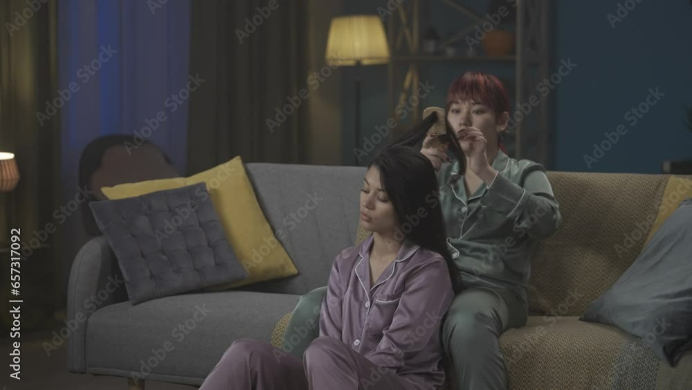 Medium-full video capturing two young women wearing pajamas sitting on the couch, gossiping, talking about something as one of them is braiding the other one's hair. Medium-full video capturing two young women wearing pajamas sitting on the couch, gossiping, talking about something as one of them is braiding the other one's hair.