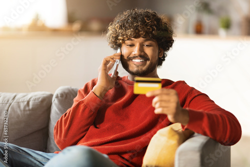 Online Banking. Smiling Young Indian Man Talking On Cellphone And Holding Credit Card