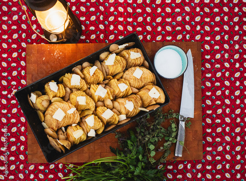 scalloped potatoes patted with butter in a steel pan on a wood cutting board. Nearby are herbs, salt, a knife, and a rustic gas lantern, all atop a red patterned tablecloth photo