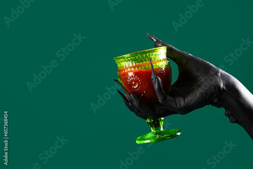 Black hand of witch with blood in glass on green background. Halloween celebration