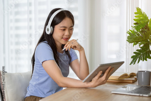 A pretty Asian woman listening to music through her headphones while using her tablet at a table.