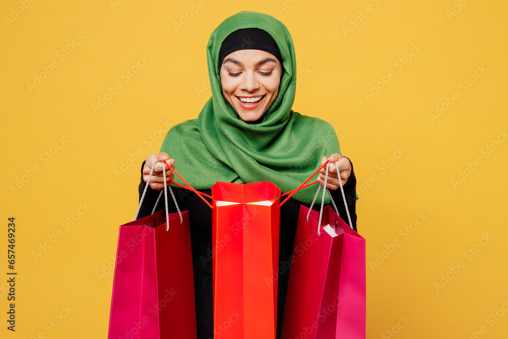 Young shocked excited excited arab asian muslim woman wears green hijab abaya clothes hold open shopping package bag isolated on plain yellow background. Black Friday sale buy day, uae islam concept.