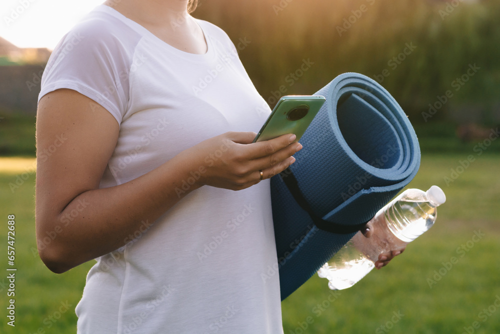 An attractive girl holds a bottle of water, a sports mat in her hands. The concept of outdoor sports in the evening. An attractive girl holds a bottle of water, a sports mat in her hands. The concept of outdoor sports in the evening.