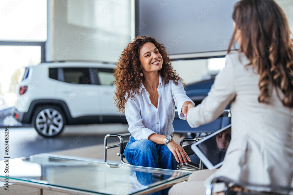 Happy woman buying a car and closing the deal with a handshake with the saleswoman at the dealership. Smiling car saleswoman discussing a contract with a female customer. - obrazy, fototapety, plakaty 
