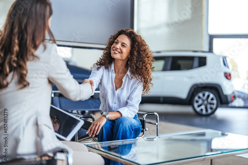 Fototapeta Happy woman buying a car and closing the deal with a handshake with the saleswoman at the dealership