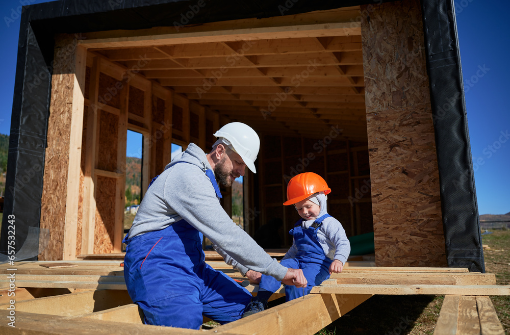 Father with toddler son building wooden frame house. Male builder playing with kid on construction site, wearing helmet and blue overalls on sunny day. Carpentry and family concept. Father with toddler son building wooden frame house. Male builder playing with kid on construction site, wearing helmet and blue overalls on sunny day. Carpentry and family concept.