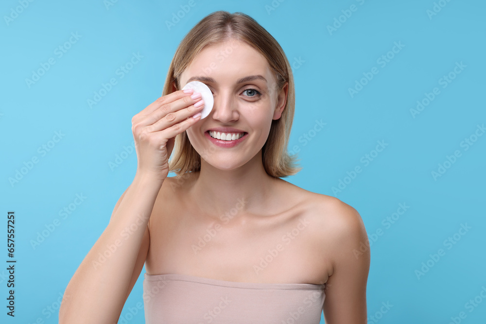 Young woman cleaning face with cotton pad on light blue background Young woman cleaning face with cotton pad on light blue background