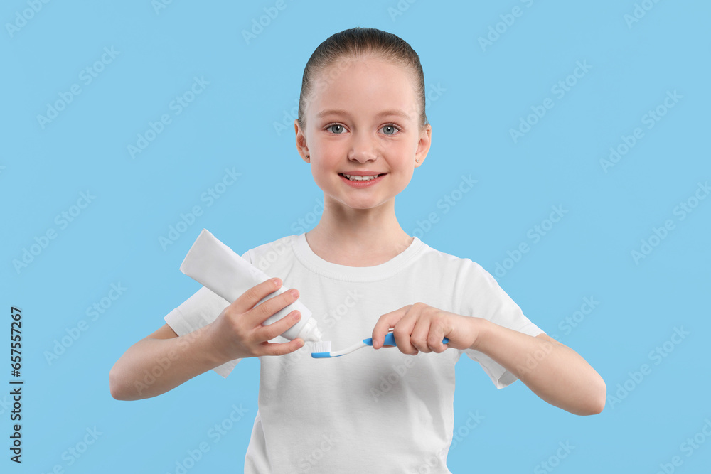Happy girl squeezing toothpaste from tube onto toothbrush on light blue background Happy girl squeezing toothpaste from tube onto toothbrush on light blue background