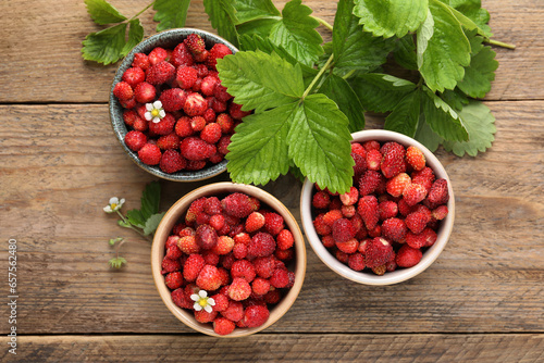 Fresh wild strawberries in bowls and leaves on wooden table, flat lay