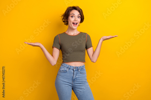 Photo of positive astonished cute woman wear trendy t-shirt palms comparing products empty space isolated on yellow color background