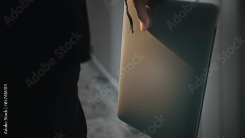 Close-up of a laptop in the hand of a man walking down a corridor. The computer is backlit, in the background is a carpeted floor. Dimmed light, modern interior. Camera in motion photo