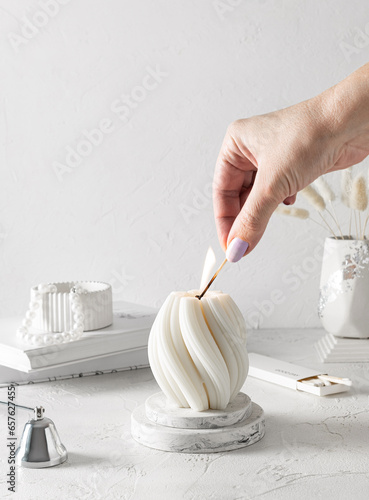 A hand with a match lights a white candle that stands on a plaster stand. A vase with white beads in the background. Matches and extinguisher on the table. White background