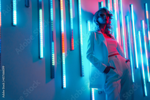 Young beautiful hipster woman in street style fashion concept. Hot model wearing white suit and sunglasses. Female posing in pink and blue neon light in studio interior