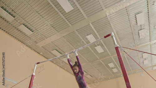 Acrobatic little girl in purple leotard practicing artistic elements on uneven bars in spacious gymnastics studio photo