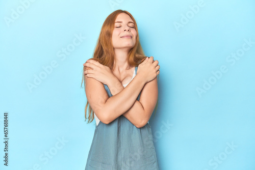 Redhead young woman on blue background hugs, smiling carefree and happy. © Asier
