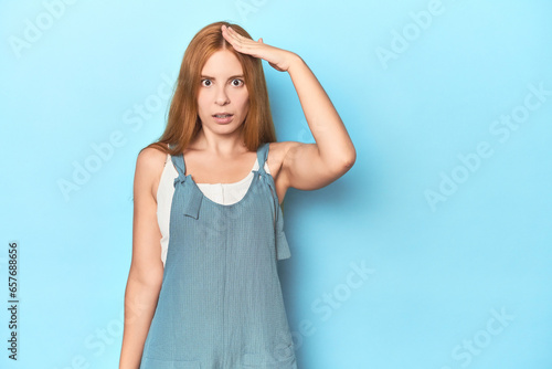 Redhead young woman on blue background shouts loud, keeps eyes opened and hands tense.