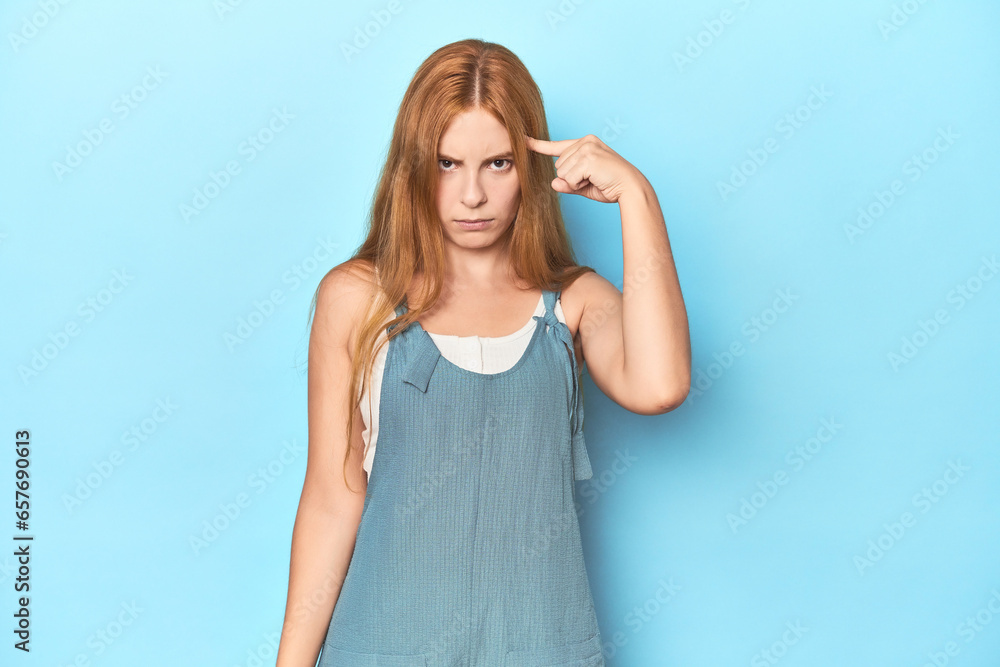 Redhead young woman on blue background pointing temple with finger, thinking, focused on a task. Redhead young woman on blue background pointing temple with finger, thinking, focused on a task.