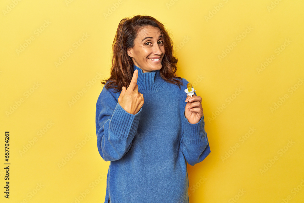 Pregnant woman with pacifier on yellow studio pointing with finger at you as if inviting come closer. Pregnant woman with pacifier on yellow studio pointing with finger at you as if inviting come closer.