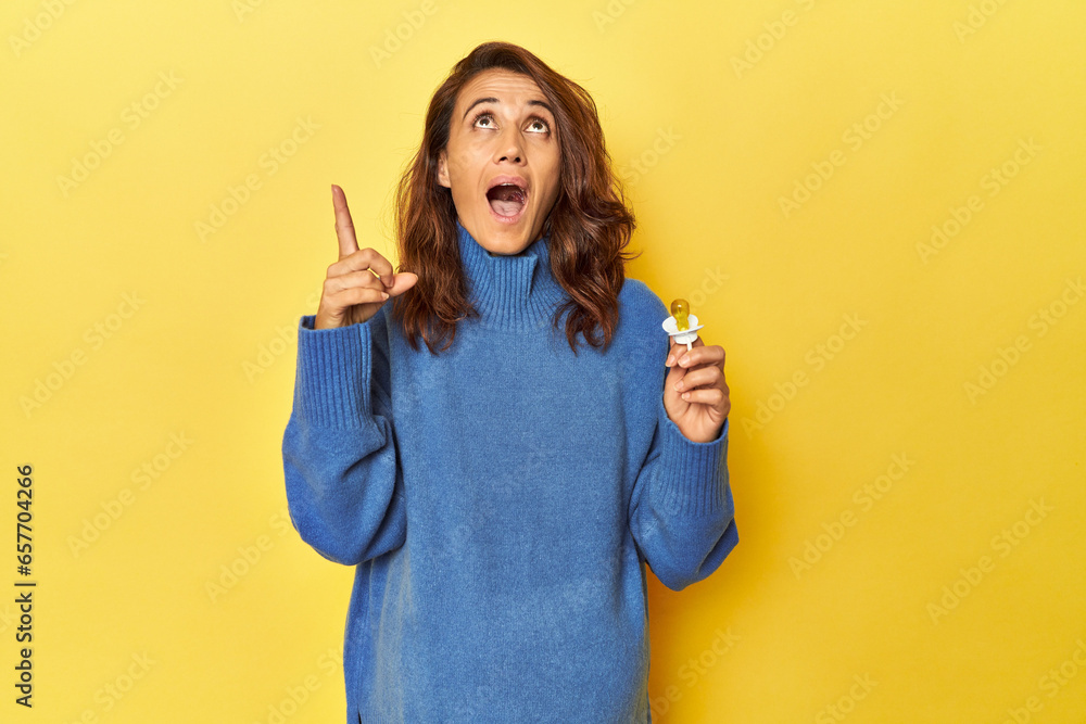 Pregnant woman with pacifier on yellow studio pointing upside with opened mouth. Pregnant woman with pacifier on yellow studio pointing upside with opened mouth.