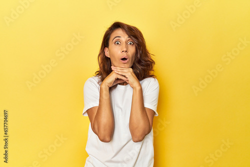 Middle-aged woman on a yellow backdrop praying for luck, amazed and opening mouth looking to front.