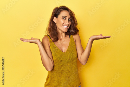 Middle-aged woman on a yellow backdrop confused and doubtful shrugging shoulders to hold a copy space.