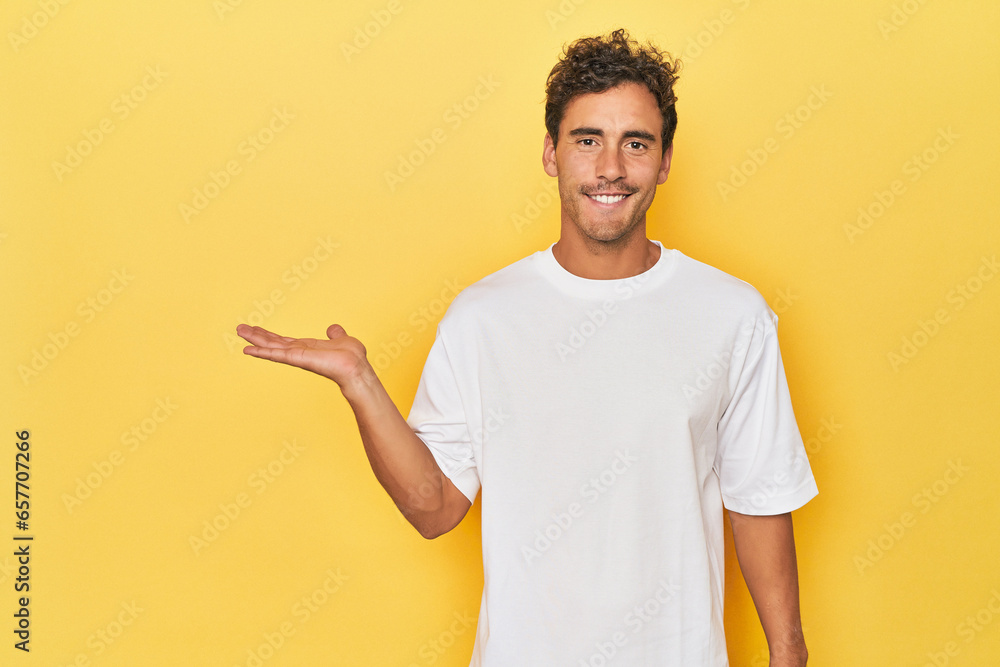 Young Latino man posing on yellow background showing a copy space on a palm and holding another hand on waist. Young Latino man posing on yellow background showing a copy space on a palm and holding another hand on waist.