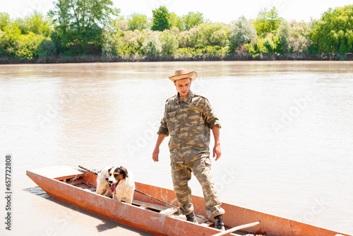 Young caucasian guy standing in shubby boat collecting tackle after fishery outdoors. Front view of male angler floating with dog, with calm, muddy river on background. Concept of fishery. photo