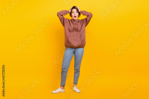 Full photo of young confused lady staring hands head denim jeans brown pullover speechless staring you isolated on yellow color background