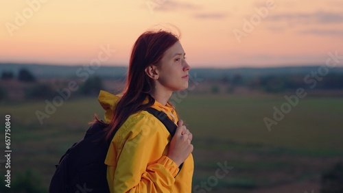 Smiling woman with backpack rests standing on large field at sunset light