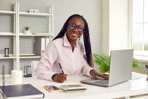 Portrait of friendly dark-skinned female financial advisor working with company accounting data. Woman is sitting at her workplace in front of laptop and calculator and is smiling at camera. photo