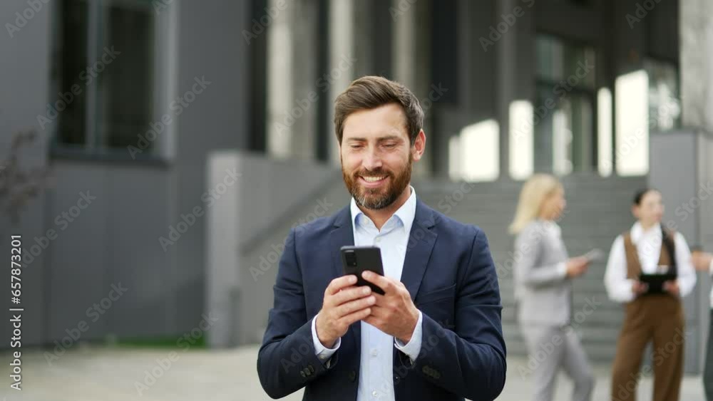 Handsome bearded business man walking near modern office building using a mobile phone. Outdoor Happy attractive male manager in formal suit go browsing chatting texting use smartphone outside. Smile Handsome bearded business man walking near modern office building using a mobile phone. Outdoor Happy attractive male manager in formal suit go browsing chatting texting use smartphone outside. Smile