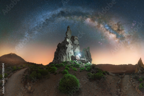 The arch of the Milky Way illuminates Los Roques de Garcia. A man with a lantern next to a gorgeous rock formation at Teide National Park in Tenerife, Canary Islands, Spain