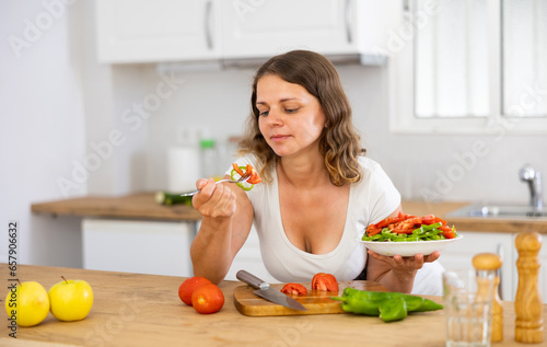 Young positive woman enjoying salad at kitchen after cooking