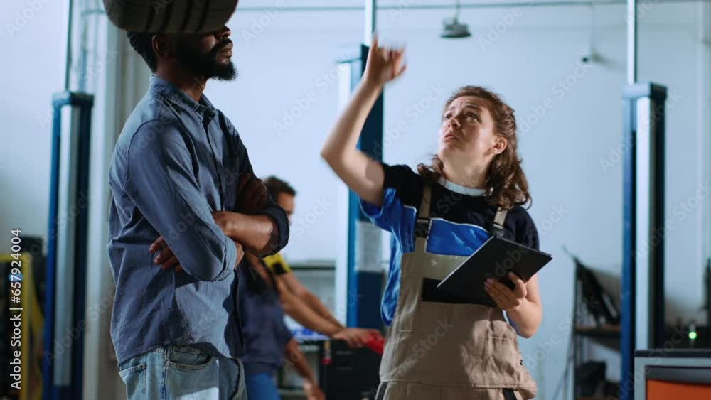 Mechanic in garage stands with customer underneath car on overhead lift, looking together for engine replacement. Expert helps BIPOC client recondition vehicle using tablet to find needed part Mechanic in garage stands with customer underneath car on overhead lift, looking together for engine replacement. Expert helps BIPOC client recondition vehicle using tablet to find needed part