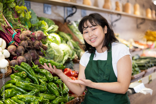 Smiling happy young asian salesgirl standing near shelves with organic fruits and vegetables in greengrocery, showing ripe green peppers photo