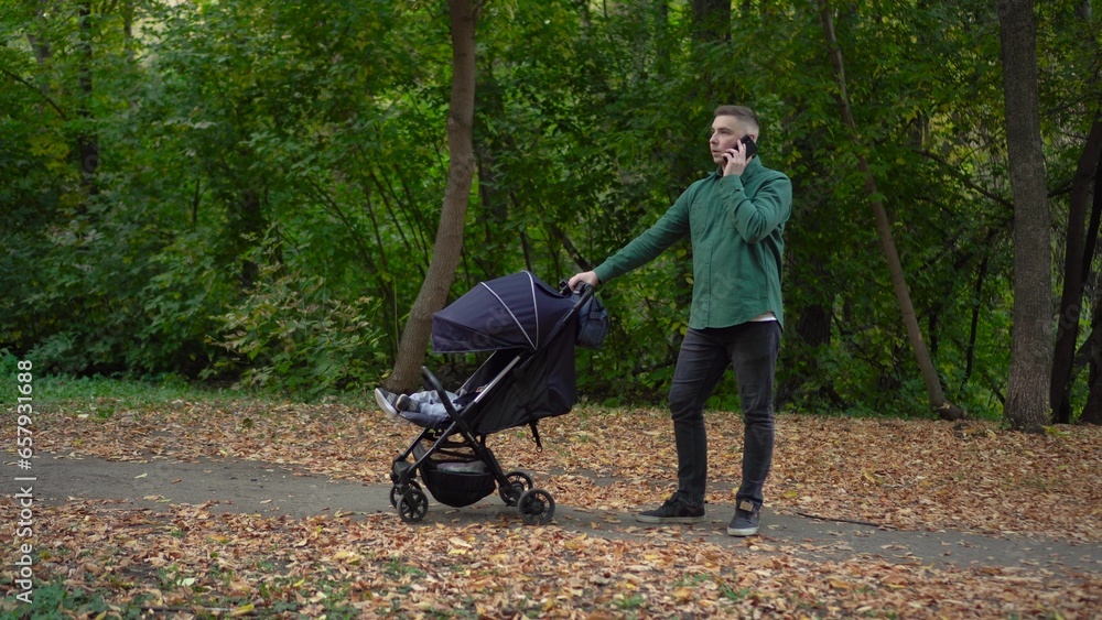 Obraz premium A young dad is talking on the phone and rocking his child in a stroller. A man speaks on the phone in the autumn forest.