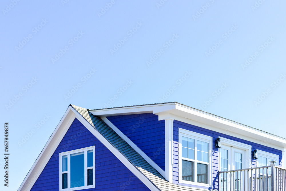The roof of a vibrant purple-colored cape cod beach house. The trim on the peaked roof is white. There are multiple glass windows in the building with a patio door and a railed patio. The sky is blue.