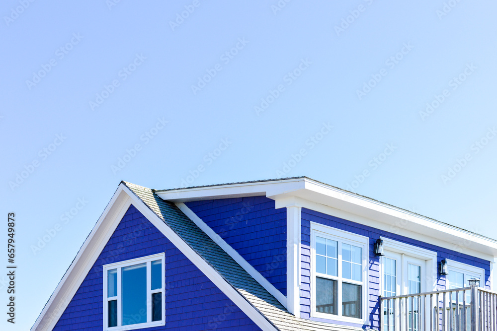 The roof of a vibrant purple-colored cape cod beach house. The trim on the peaked roof is white. There are multiple glass windows in the building with a patio door and a railed patio. The sky is blue. The roof of a vibrant purple-colored cape cod beach house. The trim on the peaked roof is white. There are multiple glass windows in the building with a patio door and a railed patio. The sky is blue.