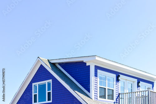 The roof of a vibrant purple-colored cape cod beach house. The trim on the peaked roof is white. There are multiple glass windows in the building with a patio door and a railed patio. The sky is blue. © Dolores  Harvey