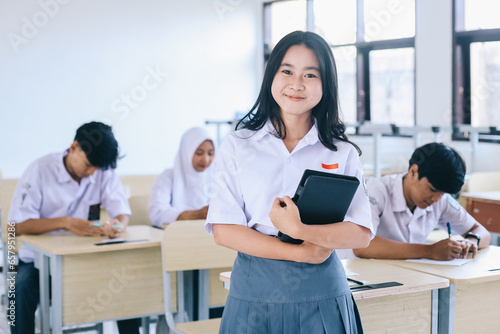 Young Asian female high school student holding a book and smiling at the camera while classmates are studying in the background © Queenmoonlite Studio
