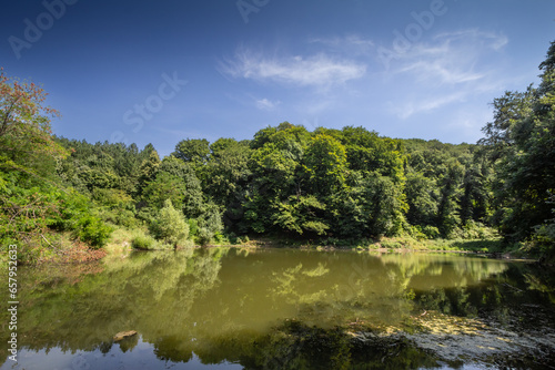 Panorama of a European natural lake, on Duboki Potok jezero lake, by Barajevo, in the southern rural part of Belgrade, Serbia, during a sunny afternoon, surrounded by a green forest.
