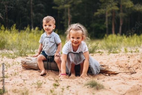 A little boy and a little girl are sitting on a log and playing with the sand on a summer day in a forest