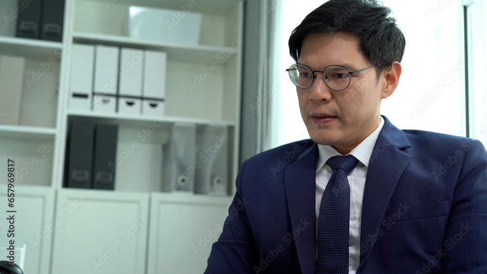 serious asian business man in suit using computer on desk disturb by mobile phone he is upset and busy in office hands on forehead serious asian business man in suit using computer on desk disturb by mobile phone he is upset and busy in office hands on forehead