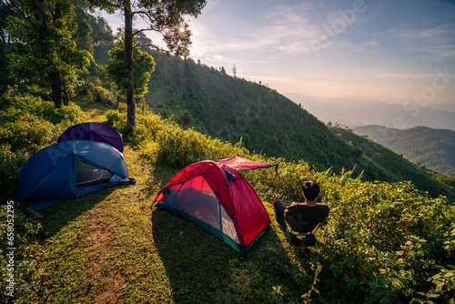 A man sitting and admiring the view with two tents in in jungle with mountain range as background in the morning (Tak province, Thailand)