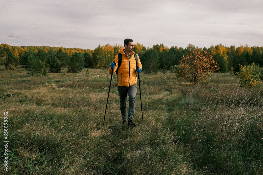 active lifestyle.A man with a backpack and hiking sticks walks through a field near the forest in autumn
